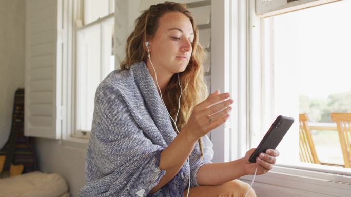 快乐的白人妇女戴着耳机坐在阳光明媚的小屋客厅使用智能手机