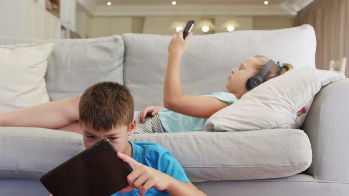 白种人兄妹用平板电脑和智能手机躺在沙发上的照片