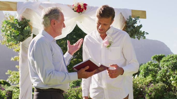 面带微笑的白人男性婚礼司仪手持书，新郎站在室外的祭坛上