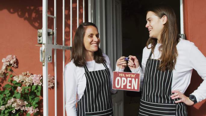 两个穿着围裙，面带微笑的白人女服务员站在门口，拿着打开的牌子