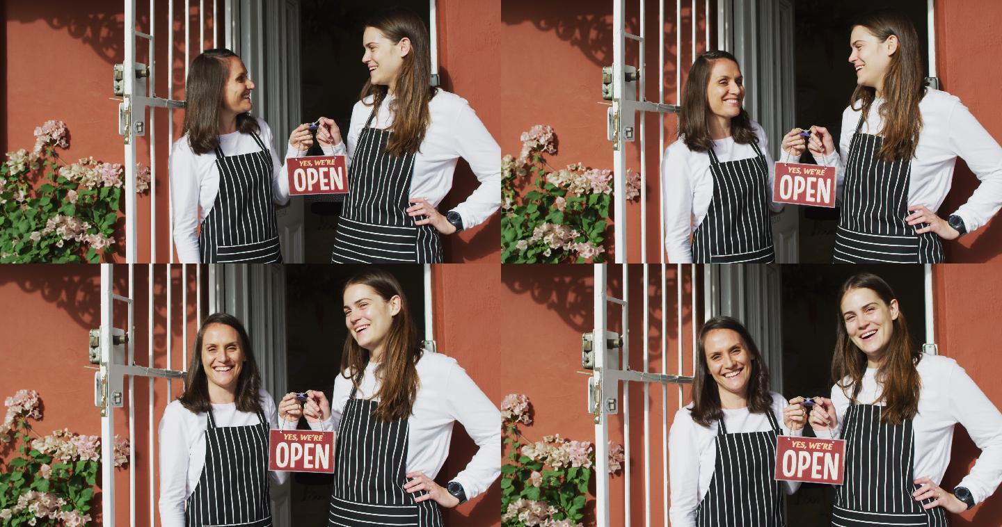 两个穿着围裙，面带微笑的白人女服务员站在门口，拿着打开的牌子