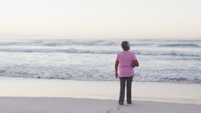 非裔美国高级妇女持有瑜伽垫看着海浪，而站在海滩上
