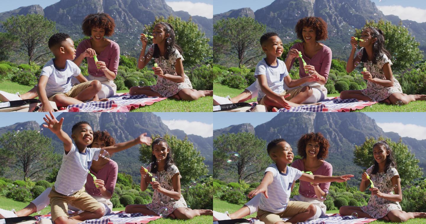 快乐的非裔美国母亲，女儿和儿子坐在户外的毯子上，吹泡泡