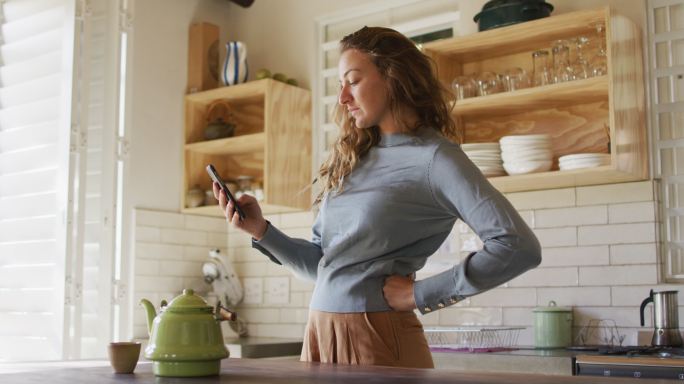白人妇女站在小屋厨房用智能手机，茶壶在柜台上