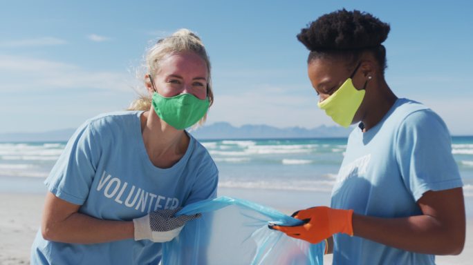两个不同的妇女穿着志愿者t恤，戴着口罩从海滩上捡垃圾