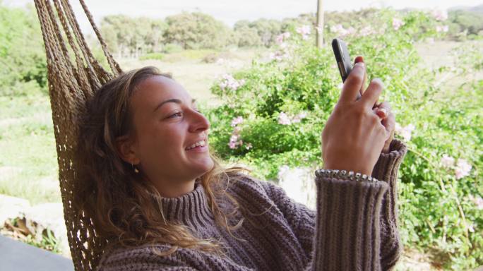 微笑的白人妇女躺在吊床上使用智能手机在阳光明媚的乡村花园