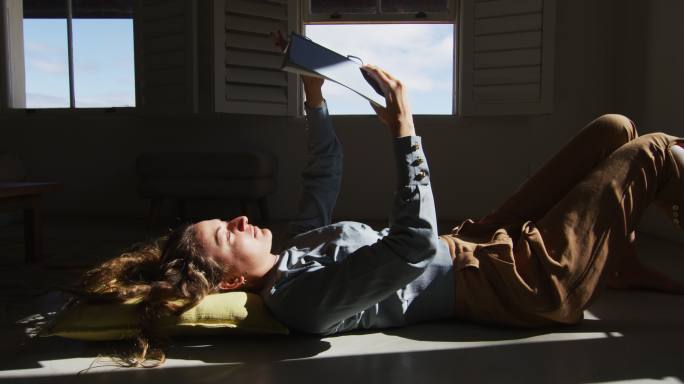 白人妇女躺在地板上拿着书和阅读在阳光明媚的小屋的客厅