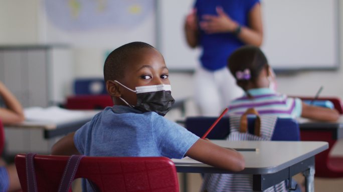 非裔美国男生的肖像坐在教室里，戴着口罩做笔记