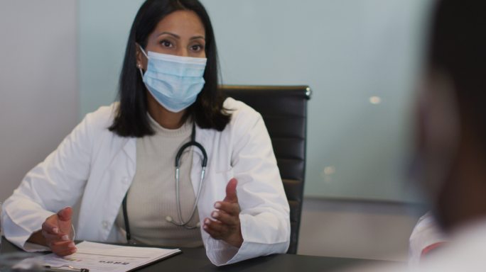 混血儿医生戴着口罩坐在会议室的桌子旁讲话