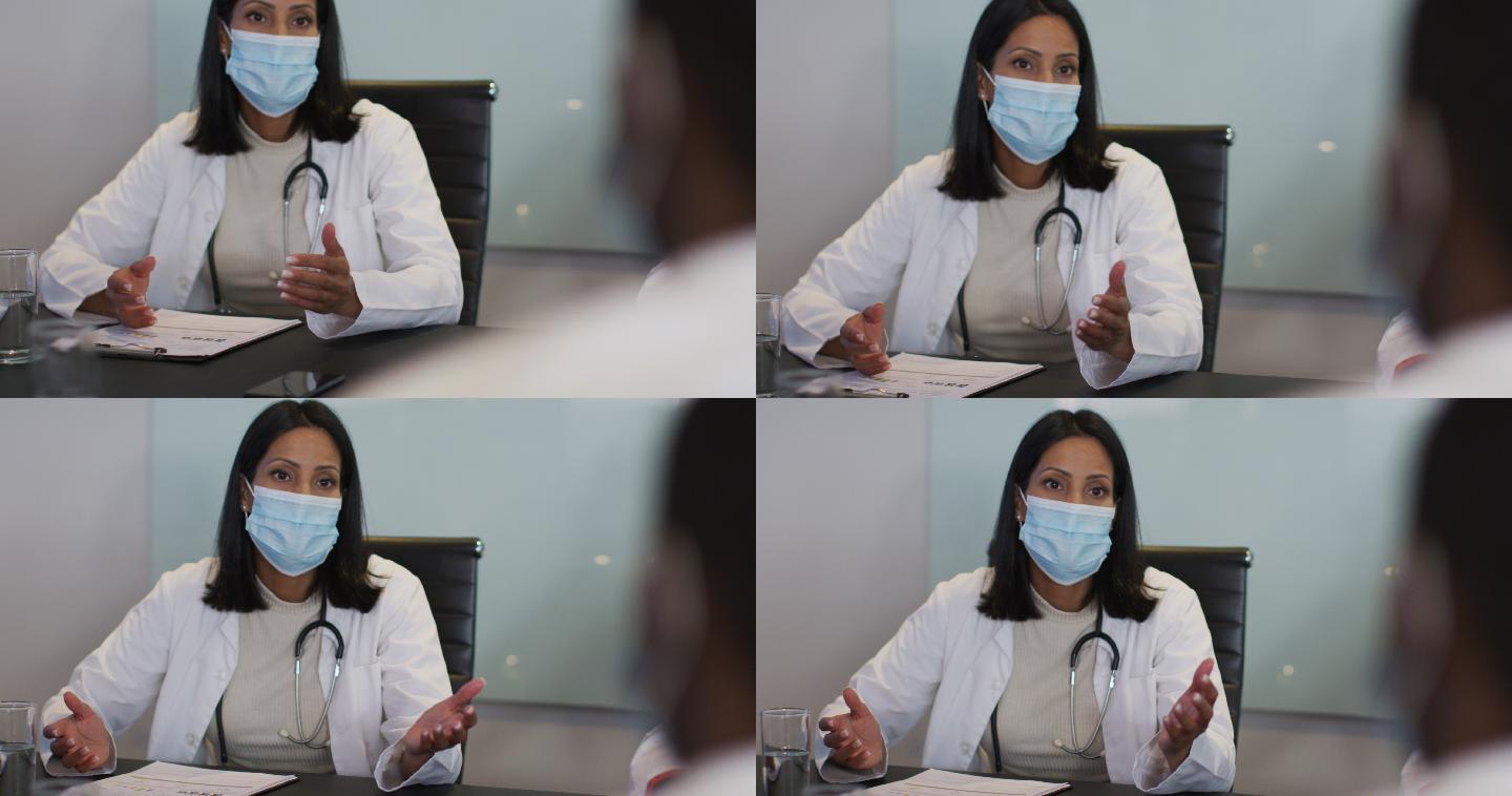 混血儿医生戴着口罩坐在会议室的桌子旁讲话