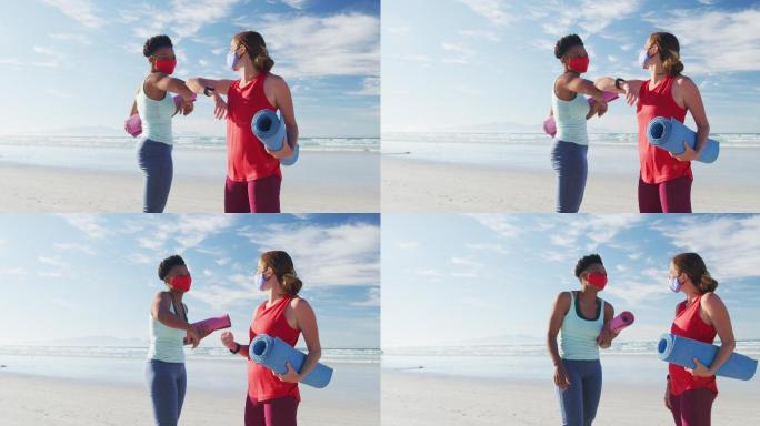 两个不同的女性朋友戴着口罩，拿着瑜伽垫，在海滩上手肘触碰