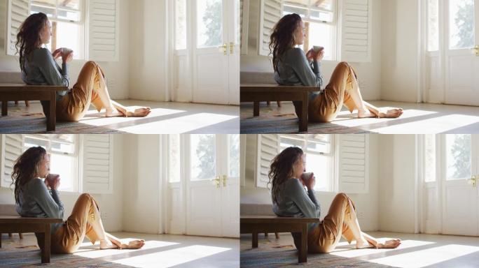 一个沉思的白人妇女坐在阳光明媚的小屋的地板上喝着咖啡，望着窗外