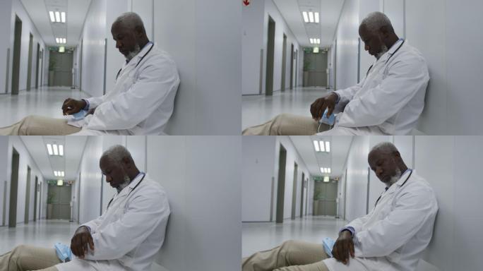 疲惫的非洲裔美国男医生坐在医院走廊上拿着口罩