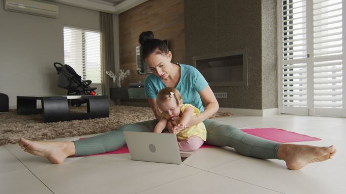 一位白人母亲抱着她的孩子在瑜伽垫上练习瑜伽，坐在家里的笔记本电脑前