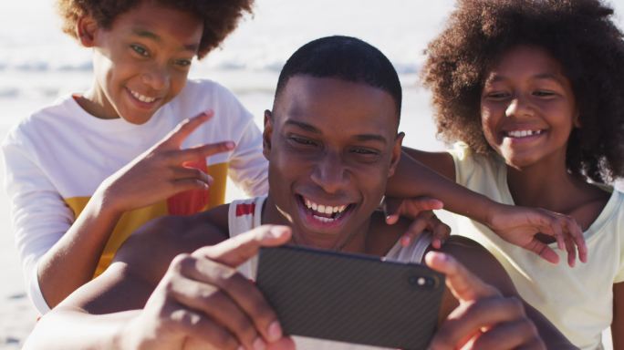 一位非洲裔美国父亲和他的孩子们在海滩上用智能手机自拍