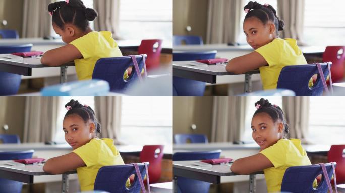 肖像快乐的混血女学生坐在教室，做笔记，看着相机