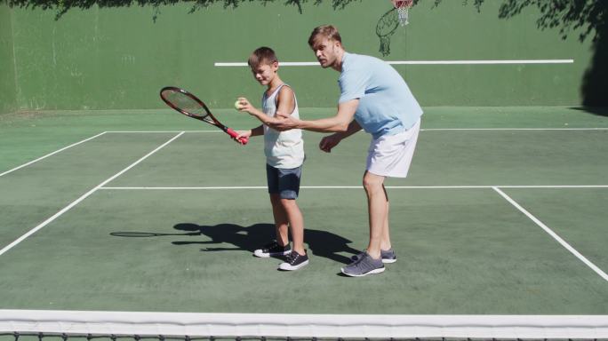一个白人父亲在一个阳光明媚的日子在网球场教他的儿子打网球