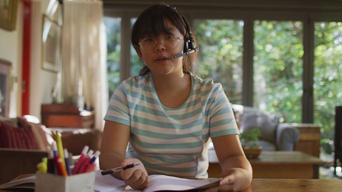 亚洲妇女坐在餐桌上戴着耳机，并进行视频通话