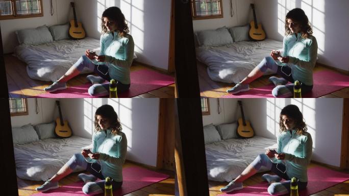 快乐的混血女子准备锻炼，使用智能手机坐在地板上，在阳光充足的卧室