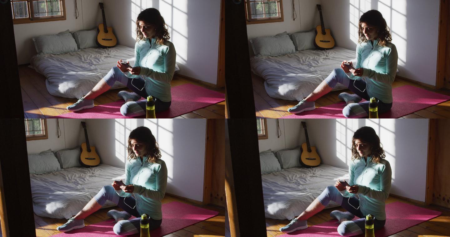快乐的混血女子准备锻炼，使用智能手机坐在地板上，在阳光充足的卧室