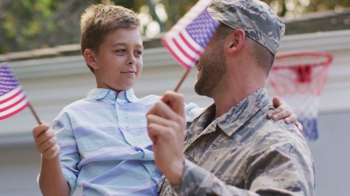 快乐的白人男性士兵带着他微笑的儿子在花园外举着旗帜
