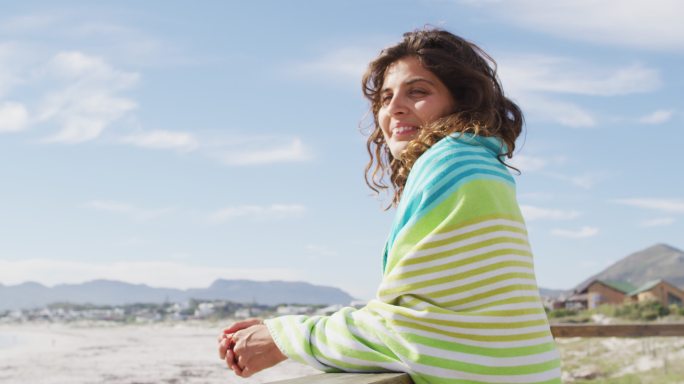 幸福的混血妇女站在海边，肩上盖着毯子，享受着风景，微笑着