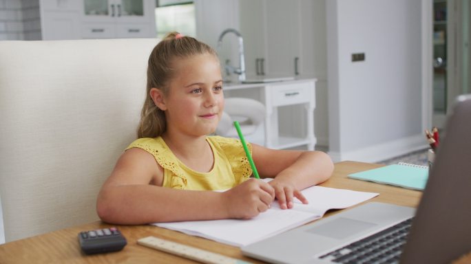 快乐的白人女孩在家里，坐在书桌上写在网上学校课程使用笔记本电脑