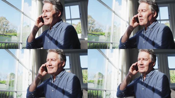 快乐的白人老人坐在阳光明媚的客厅里，用智能手机聊天，微笑着