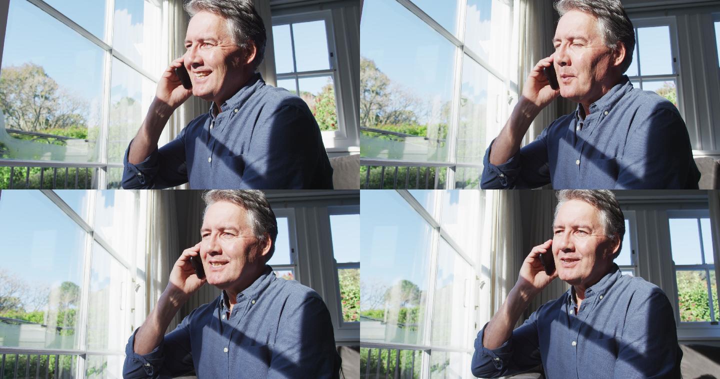 快乐的白人老人坐在阳光明媚的客厅里，用智能手机聊天，微笑着