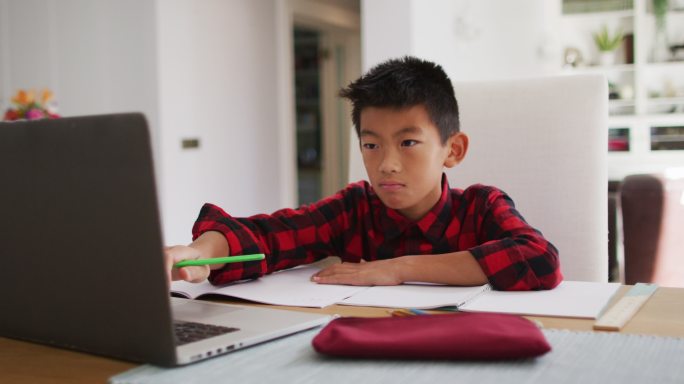 亚洲男孩在家，坐在桌旁使用笔记本电脑为在线学校课程