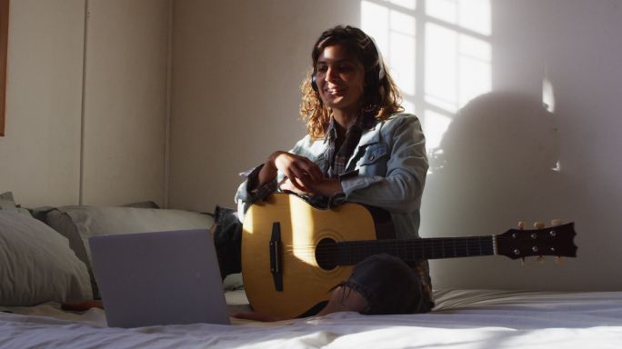 轻松的混合种族妇女耳机坐在与吉他使用笔记本电脑在阳光明媚的小屋卧室