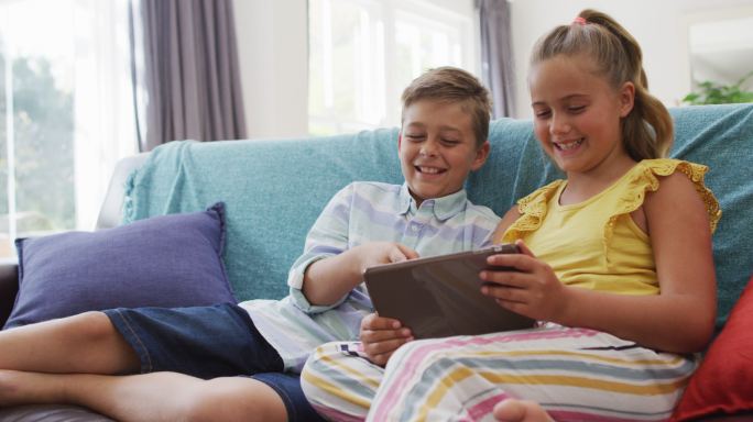 快乐的白种人兄妹在家，坐在沙发上一起使用平板电脑和微笑