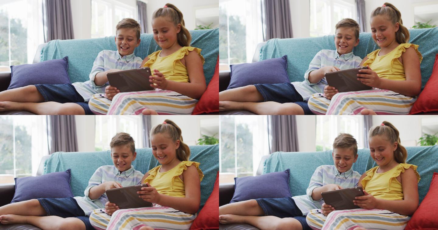 快乐的白种人兄妹在家，坐在沙发上一起使用平板电脑和微笑
