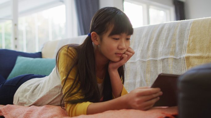 亚洲女孩微笑着躺在家里的沙发上使用平板电脑