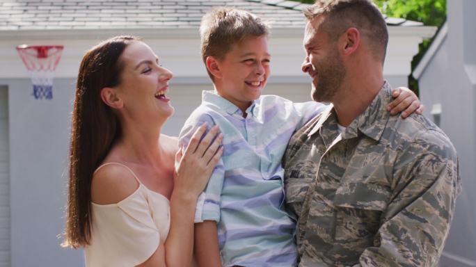 快乐的白人男性士兵拥抱他微笑的儿子和妻子在花园外的房子