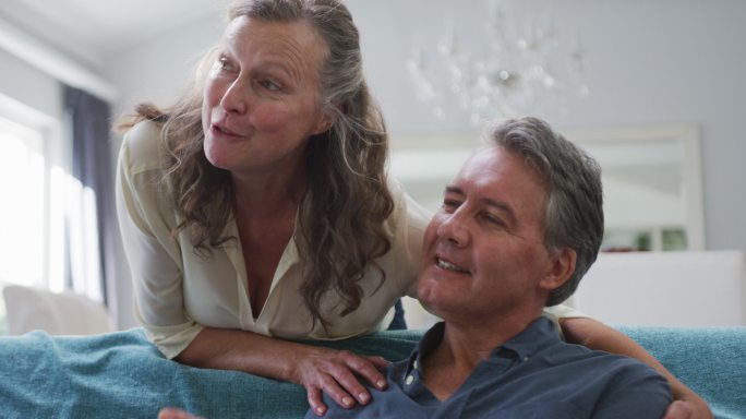 快乐的白种人夫妇在客厅看电视和聊天，妻子拥抱坐着的丈夫