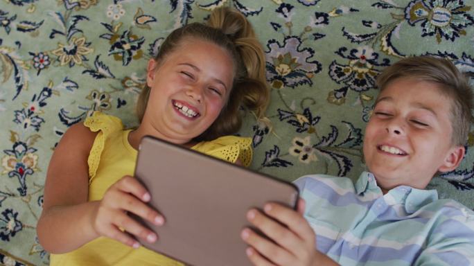 快乐的白种人兄妹在家，一起躺在地板上用平板电脑，笑着