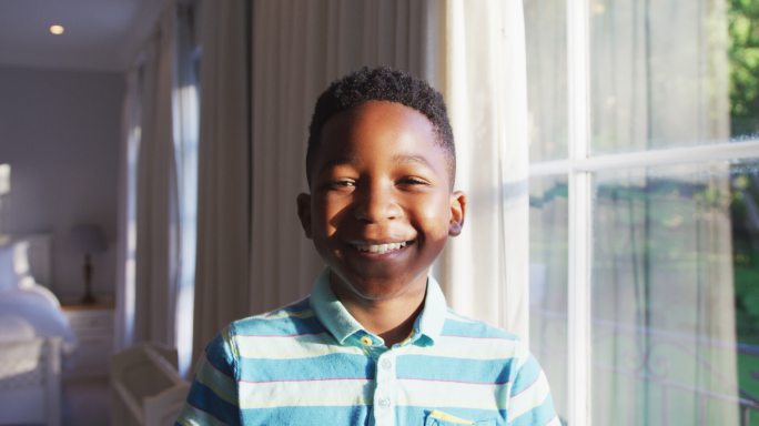 快乐的非裔美国男孩的肖像，望着阳光明媚的窗户，转向相机和微笑