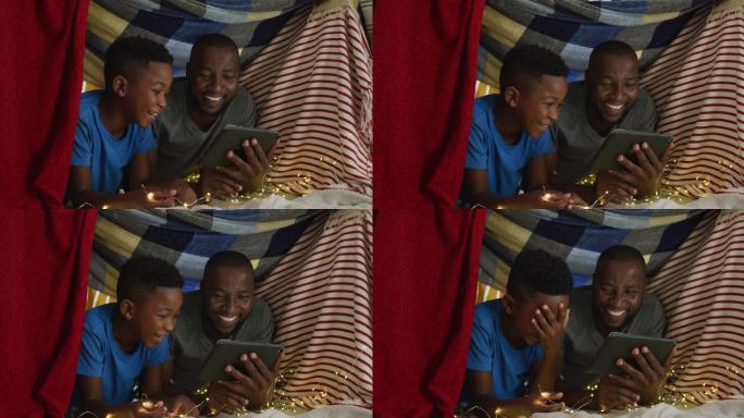 快乐的非洲裔美国父子使用平板电脑和大笑躺在毯子营地