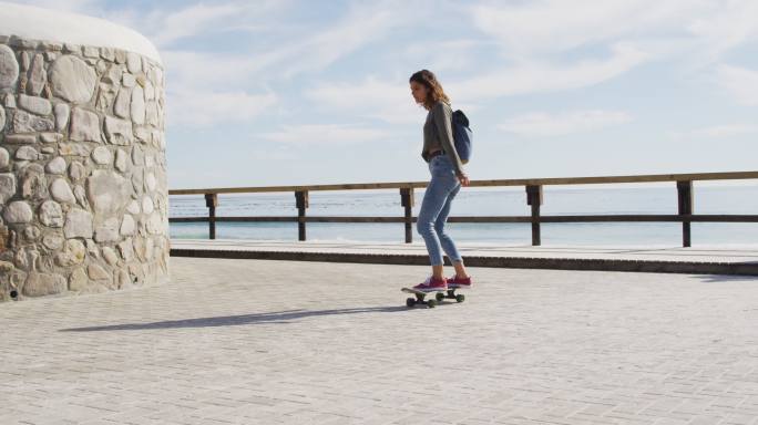 快乐的混合种族妇女滑板在阳光明媚的海滨长廊