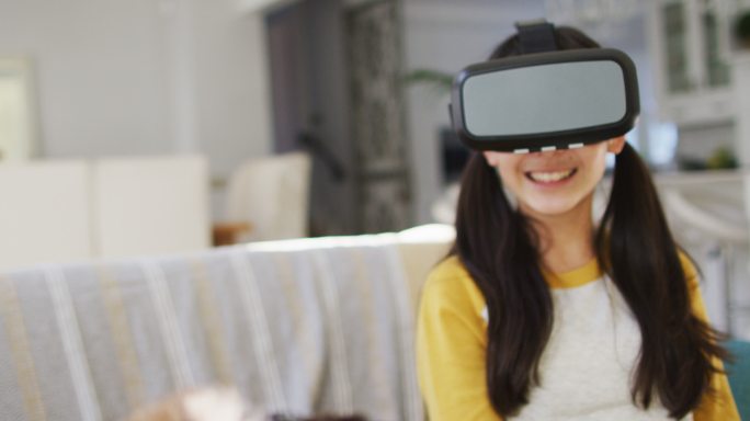 快乐的亚洲兄弟和姐妹在家，坐在沙发上在客厅使用虚拟现实头盔的乐趣