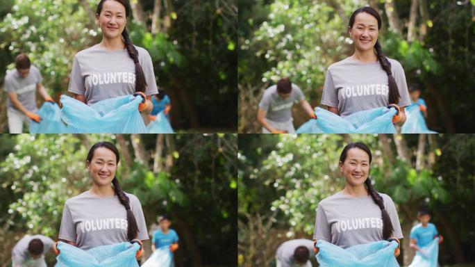 微笑的亚洲妇女穿着志愿者t恤，拿着垃圾袋收集塑料垃圾