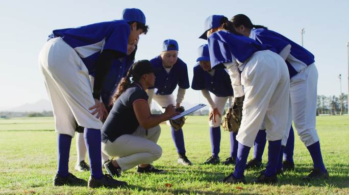 各种各样的女棒球运动员挤在女教练周围，蹲着，在球场上聊天