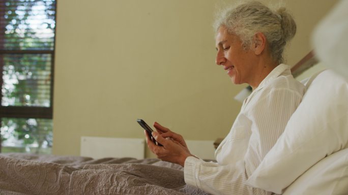 高级混血妇女躺在床上用智能手机和微笑