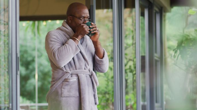 非裔美国老人穿着浴袍站在阳台上喝着咖啡，欣赏风景