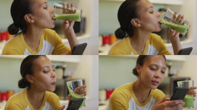混血儿妇女喝着健康的饮料，在厨房里使用智能手机
