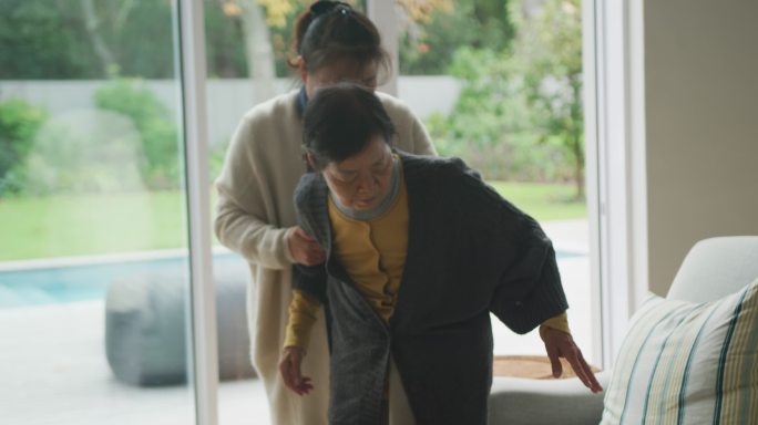 年长的亚洲母亲在客厅的成年女儿帮助下坐在扶手椅上