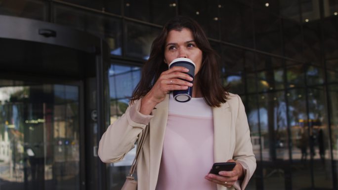 白人女商人在现代办公室外使用智能手机和喝外卖咖啡