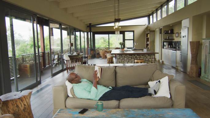 快乐的非洲裔美国老人放松在客厅，躺在沙发上看书