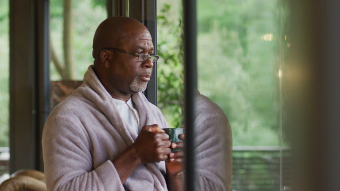 深思熟虑的非裔美国老人穿着浴袍，喝着咖啡，看着窗外